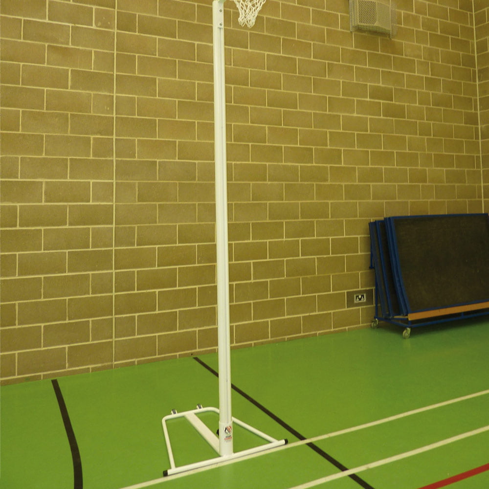 A portable netball hoop with a white pole and base stands on a green indoor court against a tan brick wall. Folded blue mats are stacked against the wall in the background.