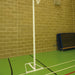 A portable netball hoop with a white pole and base stands on a green indoor court against a tan brick wall. Folded blue mats are stacked against the wall in the background.