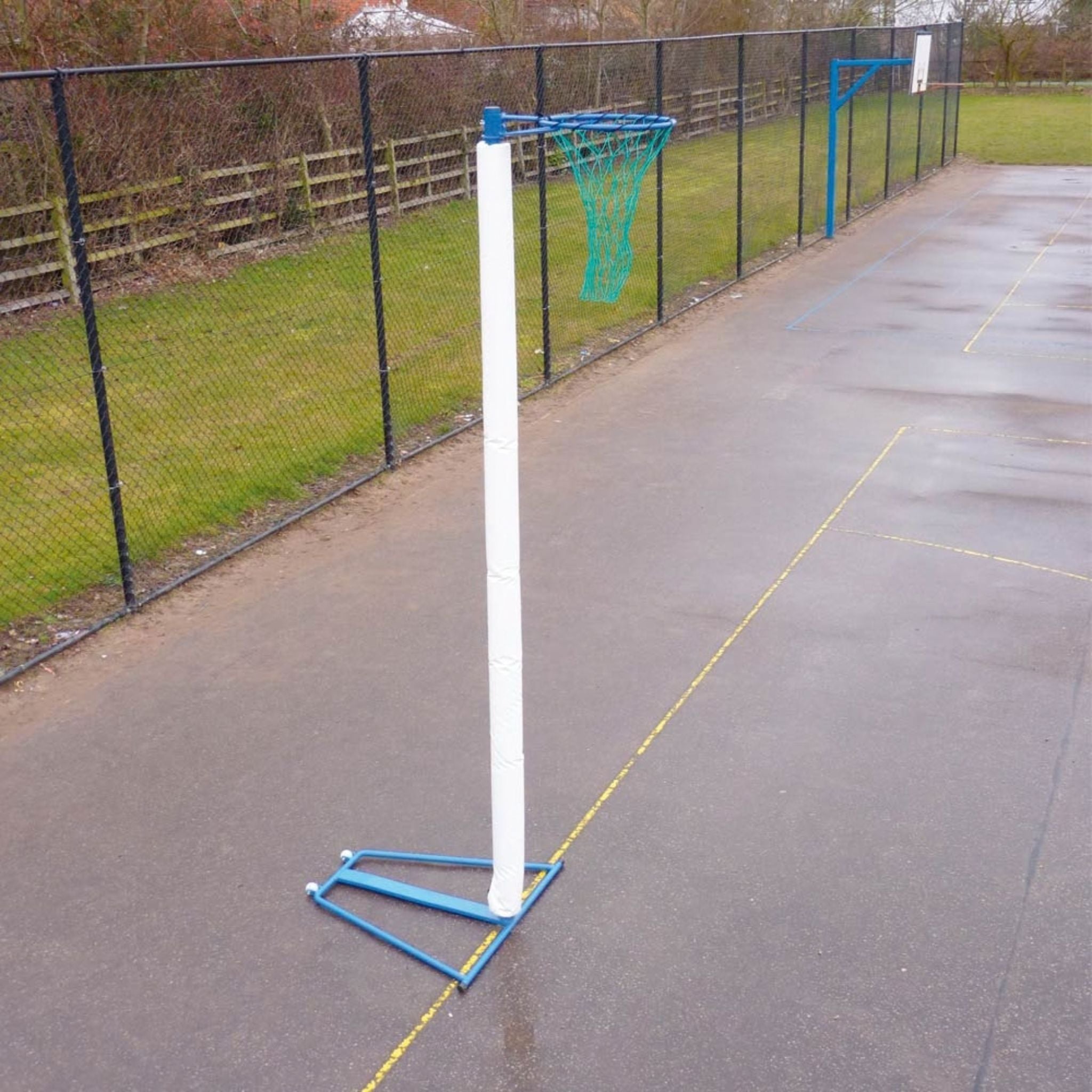 A portable netball hoop with a white padded pole, blue base, and green net, set up on an outdoor paved court next to a chain-link fence.