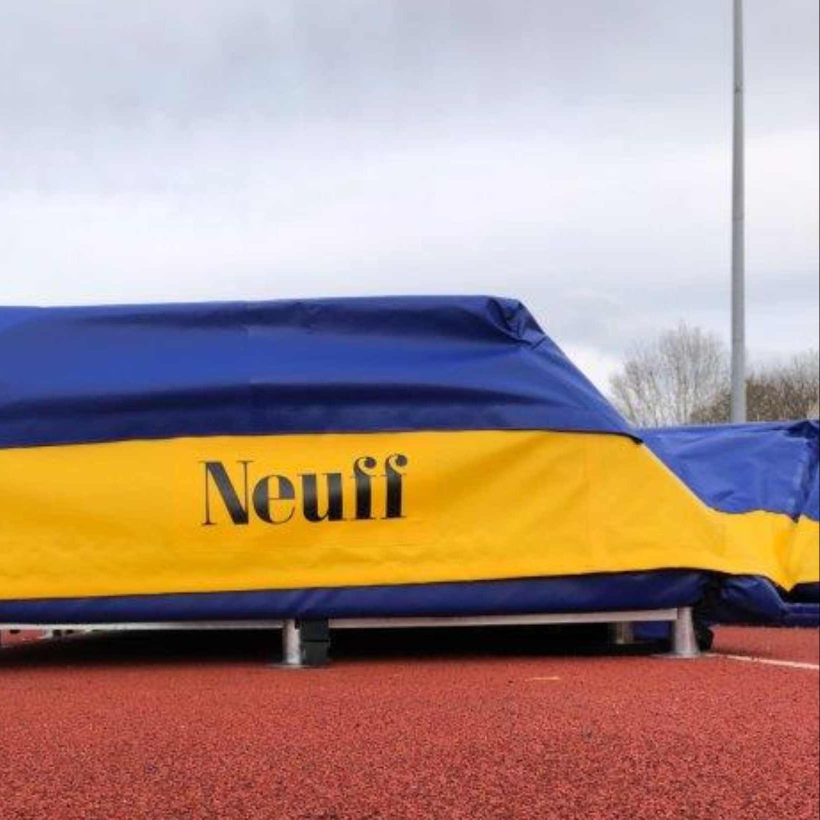 A blue and yellow PVC cover with the brand logo 'Neuff' on a landing bed for pole vault or high jump, placed on a track field.