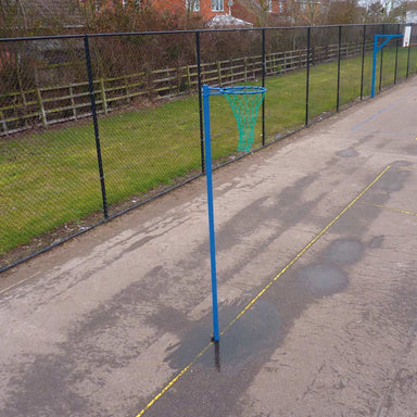 A blue socketed netball hoop with a green net on an outdoor paved court, with a chain-link fence and grass in the background.