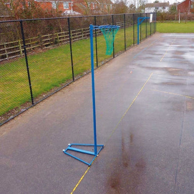 A blue portable netball hoop with a green net stands on a wet paved outdoor court next to a black chain-link fence.