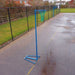 A blue portable netball hoop with a green net stands on a wet paved outdoor court next to a black chain-link fence.