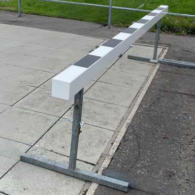 White wooden steeplechase barrier with metal adjustable legs on a concrete surface with grass in the background