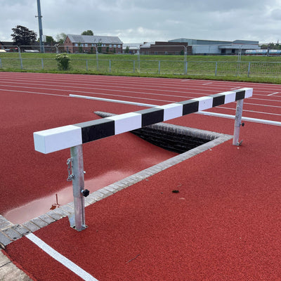 Athletic steeplechase hurdle next to an empty water trough on a running track with buildings and trees in the background
