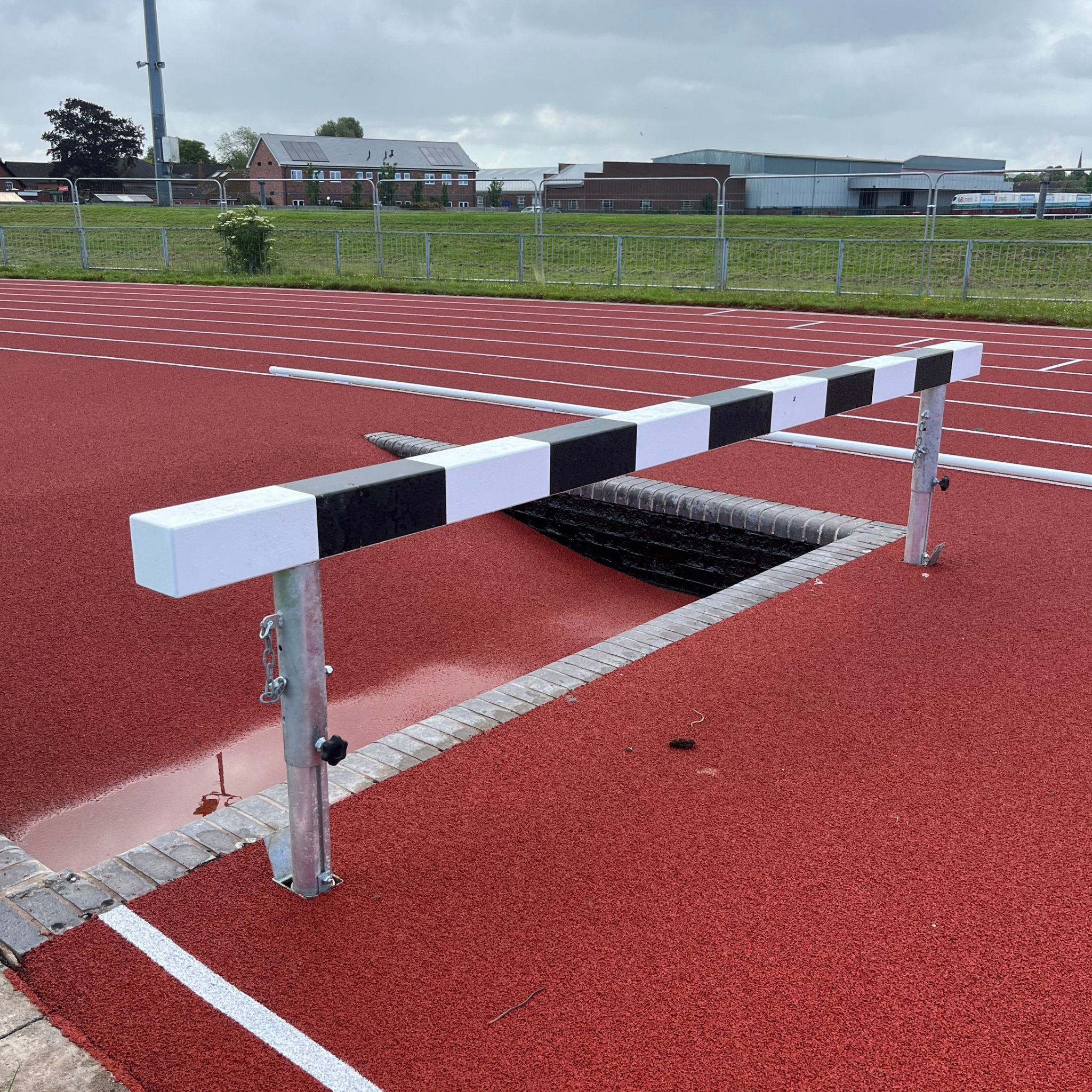 Athletic steeplechase hurdle next to an empty water trough on a running track with buildings and trees in the background