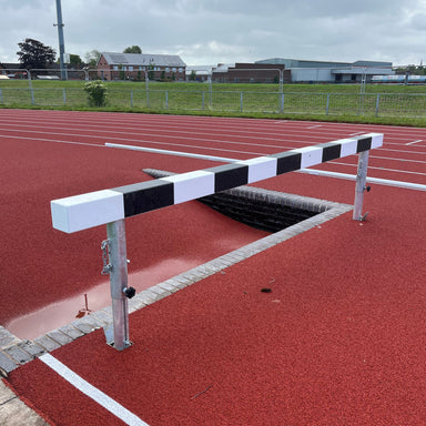 Athletic steeplechase hurdle next to an empty water trough on a running track with buildings and trees in the background