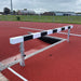 Athletic steeplechase hurdle next to an empty water trough on a running track with buildings and trees in the background